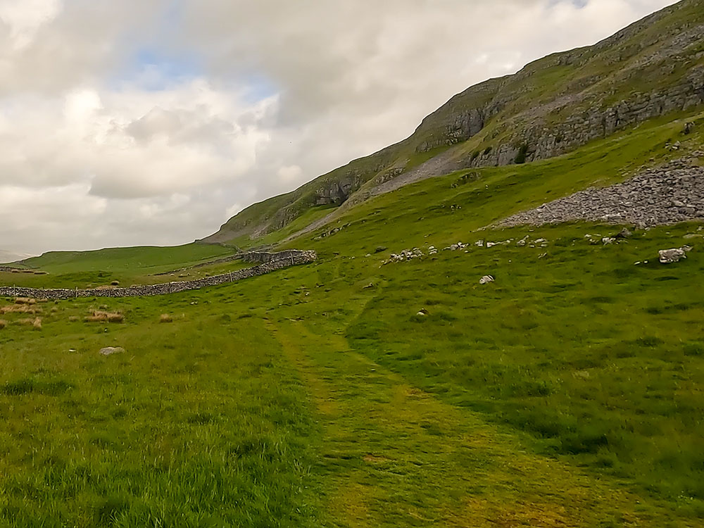 Wide grassy path that heads around to the right-hand side of the wall ahead